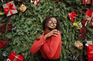 porträt einer lockigen afrikanerin mit modischem rotem rollkragenpullover, die sich gegen weihnachtsdekorationen posiert, silvesterthema. foto