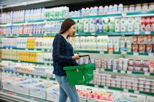 Einkaufsfrau, die die Regale im Supermarkt betrachtet. Porträt eines jungen Mädchens in einem Marktladen mit grünem Einkaufskorb und Milchproduktion. foto