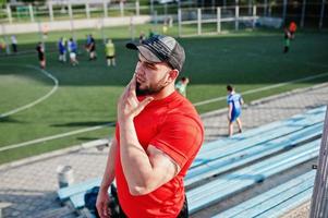 junger brutaler bärtiger muskulöser mann trägt rotes hemd, shorts und mütze im stadion. foto