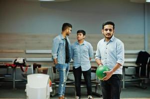 südasiatischer mann im jeanshemd, der an der kegelbahn mit ball auf händen steht. Kerl bereitet sich auf einen Wurf vor. Freunde unterstützen ihn lautstark. foto