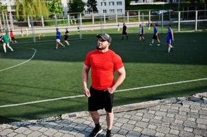 junger brutaler bärtiger muskulöser mann trägt rotes hemd, shorts und mütze im stadion. foto