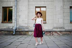 Modisches und schönes blondes Modelmädchen in stylischem rotem Samt-Veloursrock, weißer Bluse und Hut, posiert mit Telefon und Kopfhörern. foto