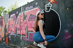 Rückseite des Gesäßes stilvolles, lässiges Hipster-Mädchen in Mütze, Sonnenbrille und Jeans, das Musik über Kopfhörer des Mobiltelefons gegen große Graffiti-Wand mit großer TNT-Bombe hört. foto