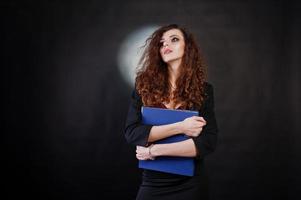 brünettes lockiges Mädchen in schwarzer Bürojacke mit Rock, auf BH mit Ordner mit Dokumenten in Händen auf Studio vor schwarzem Hintergrund. sexy Geschäftsfrau. foto