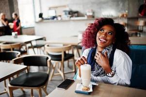 attraktives afroamerikanisches lockiges Mädchen, das im Café mit Latte sitzt. foto