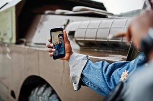 afroamerikanischer mann in jeansjacke, baskenmütze und brille, raucht zigarre und posiert gegen btr-militärpanzerfahrzeug, macht selfie am telefon. konzentrieren sie sich auf den bildschirm des handys. foto