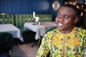 Nahaufnahme Porträt eines freundlichen Afro-Mannes in traditioneller gelber Kleidung, der im Restaurant sitzt. foto