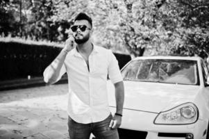 Stilvolles großes arabisches Mannmodell in weißem Hemd, Jeans und Sonnenbrille posierte auf der Straße der Stadt. bartreicher attraktiver arabischer kerl gegen weißes suv-auto, das am handy spricht. foto