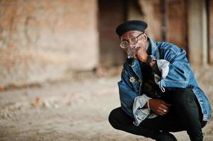 afroamerikanischer mann in jeansjacke, baskenmütze und brille, rauchende zigarre in verlassener fabrik. foto