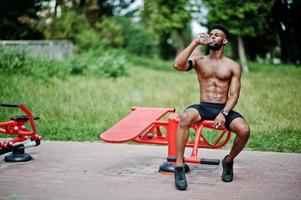 afroamerikanischer männlicher athlet sexy sport mit nacktem torso mann mit laufendem sportarmgehäuse für handy, übungen auf den straßentrainingssimulatoren und trinkwasser aus der flasche. foto