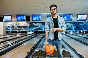 stylischer bart asiatischer mann in jeansjacke und brille, der an der bowlingbahn mit ball zur hand steht. foto
