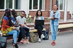 Gruppe von fünf afrikanischen College-Studenten, die gemeinsam Zeit auf dem Campus des Universitätshofs verbringen. schwarze afrofreunde, die an der bank mit schulsachen, laptops notizbüchern studieren. foto