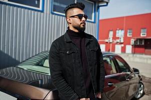 Mode arabischer Mann trägt schwarze Jeansjacke und Sonnenbrille gegen modernes Geschäftsauto. stylischer, erfolgreicher und modischer arabischer Modeltyp. foto