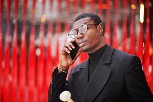 Stilvoller afroamerikanischer Gentleman in eleganter schwarzer Jacke, der einen Retro-Spazierstock als Rohrkolben oder einen Kippstock mit goldenem Diamantkugelgriff hält. reicher modischer afro-mann gegen rote wand. foto