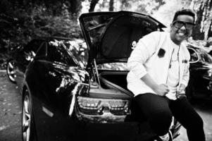 stylischer hipster arabischer mann mit sonnenbrille posierte im freien auf der straße und saß auf dem kofferraum seines schwarzen muskelautos. reicher schwarzer Mann. foto
