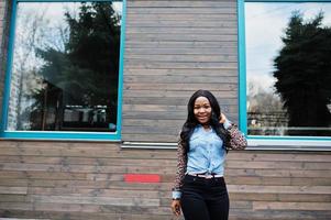 Hipster afroamerikanisches Mädchen mit Jeanshemd und Leopardenärmeln posiert auf der Straße gegen Holzhaus mit Fenstern. foto