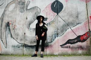 sinnliches Mädchen ganz in Schwarz, roten Lippen und Hut. gotische dramatische frau hält weiße chrysanthemenblume gegen graffitiwand. foto