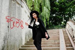 sinnliches Mädchen ganz in Schwarz, roten Lippen und Hut. gotische dramatische frau hält weiße chrysanthemenblume gegen graffitiwand. foto