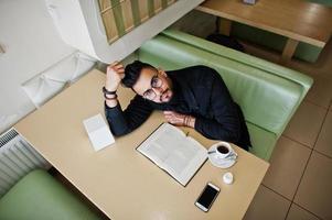 arabischer mann trägt schwarze jeansjacke und brille, sitzt im café, liest buch und trinkt kaffee. stilvoller und modischer arabischer Modelltyp. foto
