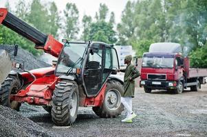 stylischer afroamerikanischer mann mit hut und sonnenbrille posierte im regen gegen traktor mit einem eimer. foto