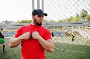 junger brutaler bärtiger muskulöser mann trägt rotes hemd, shorts und mütze im stadion. foto