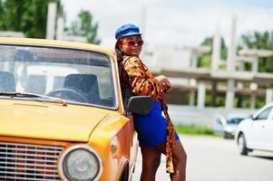 Schöne afrikanisch-amerikanische Dame mit Sonnenbrille, die in der Nähe eines orangefarbenen klassischen Retro-Autos steht. foto