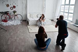 das team von zwei fotografen fotografiert zwillinge model mädchen, während sie auf die fernbedienung des fernsehers im studio klicken. professioneller fotograf bei der arbeit. foto