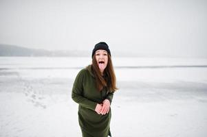 lustige mädchenkleidung auf langem grünem sweatshirt, jeans und schwarzer kopfbedeckung zeigt eine zunge am zugefrorenen see am wintertag. foto