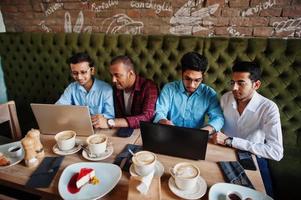 Gruppe von vier südasiatischen Männern bei Geschäftstreffen im Café gestellt. inder arbeiten mit laptops zusammen, verwenden verschiedene geräte und unterhalten sich. foto