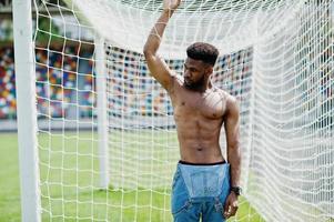 hübscher sexy afroamerikanischer muskulöser Mann mit nacktem Oberkörper in Jeans-Overalls posiert auf grünem Gras gegen Fußballtore des Stadionfeldes. modisches Porträt eines schwarzen Mannes. foto