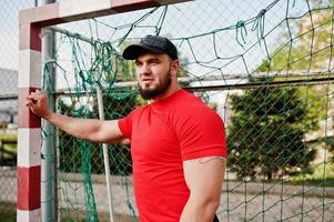 junger brutaler bärtiger muskulöser mann trägt rotes hemd, shorts und mütze im stadion. foto