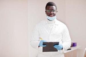 afrikanisch-amerikanischer männlicher arzt mit brille und maske posierte in der klinik mit schwarzem klemmbrett zur hand. foto