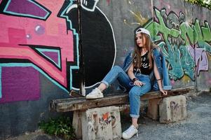 stilvolles, lässiges hipster-mädchen in mütze und jeans, das musik über kopfhörer des handys gegen große graffitiwand mit bombe hört. foto