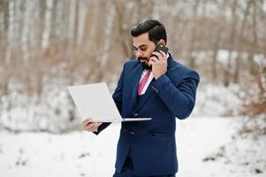 stilvoller indischer bartgeschäftsmann im anzug, der am wintertag im freien mit laptop in der hand posiert und am handy spricht. foto