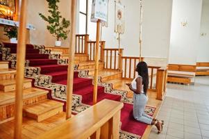 Afroamerikanerin, die in der Kirche betet. Gläubige meditieren in der Kathedrale und spirituelle Gebetszeit. Afro-Mädchen halten Rosenkranz und knien. foto