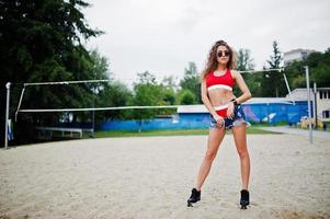 sexy lockiges Model-Mädchen in rotem Top, Jeans-Denim-Shorts, Tasse und Sonnenbrille am Beach-Volleyball-Feld. foto