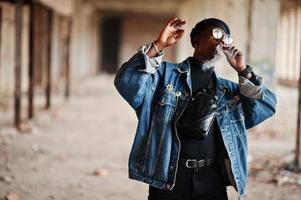 afroamerikanischer mann in jeansjacke, baskenmütze und brille, rauchende zigarre in verlassener fabrik. foto