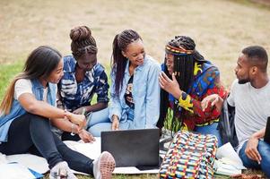 Gruppe von fünf afrikanischen College-Studenten, die gemeinsam Zeit auf dem Campus des Universitätshofs verbringen. schwarze Afro-Freunde sitzen auf Gras und lernen mit Laptops. foto