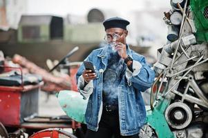 afroamerikanischer mann in jeansjacke, baskenmütze und brille, raucht zigarre und posiert gegen altes retro-fahrzeug und schaut aufs telefon. foto