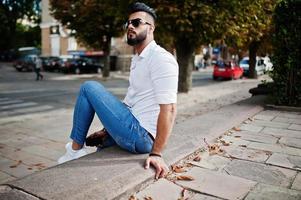 Stilvolles großes arabisches Mannmodell in weißem Hemd, Jeans und Sonnenbrille, das auf der Straße der Stadt sitzt. Bart attraktiver arabischer Typ im Park. foto