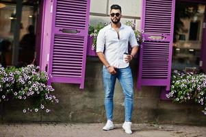 Stilvolles großes arabisches Mannmodell in weißem Hemd, Jeans und Sonnenbrille posierte auf der Straße der Stadt. bart attraktiver arabischer kerl mit tasse kaffee gegen lila fenster. foto
