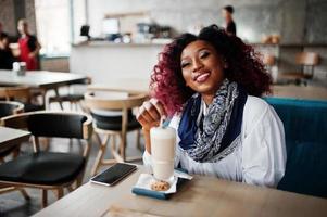 attraktives afroamerikanisches lockiges Mädchen, das im Café mit Latte sitzt. foto