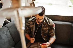 stylischer bart arabischer mann in brille und militärjacke raucht shisha an der straßenbar. Arabisches Model, das sich ausruht und am Telefon schaut. foto