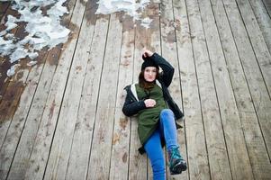 junge Mädchen tragen lange grüne Sweatshirts, Jeans und schwarze Kopfbedeckungen, die auf Holzboden mit Schnee sitzen. foto