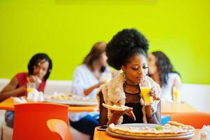 afrikanische frau mit pizza sitzt im restaurant und trinkt saft gegen dunkelhäutige mädchen. foto