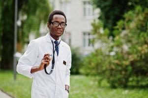 stilvoller afroamerikanischer arzt mit stethoskop und laborkittel, bei brille im freien gestellt. foto