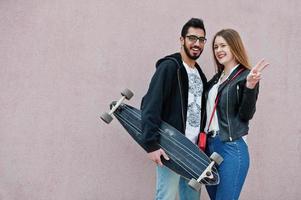 cooles gemischtrassiges paar, das mit longboard gegen rosa wand posiert. foto