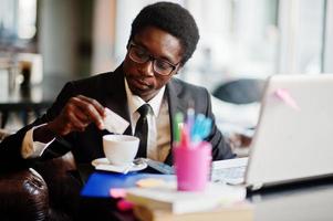 Business-Afroamerikaner trägt schwarzen Anzug und Brille, sitzt im Büro mit Laptop und arbeitet, gießt Zucker in Kaffee. foto