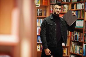 großer, intelligenter arabischer student, trägt schwarze jeansjacke und brille, in der bibliothek mit buch zur hand. foto