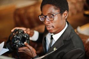 afroamerikanischer fotograf paparazzi mann trägt schwarzen anzug und brille, sitzt mit kamera im büro und arbeitet hinter laptop. foto